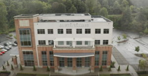 Aerial view of a four-story brick and white building with "South College" on the facade. The building is surrounded by a parking lot and trees. The weather appears overcast.